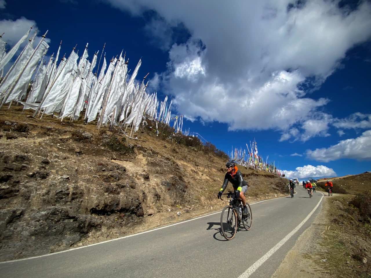 A cyclist on a tour in Bhutan crests the Chela La and passes white prayer flags beneath a blue sky