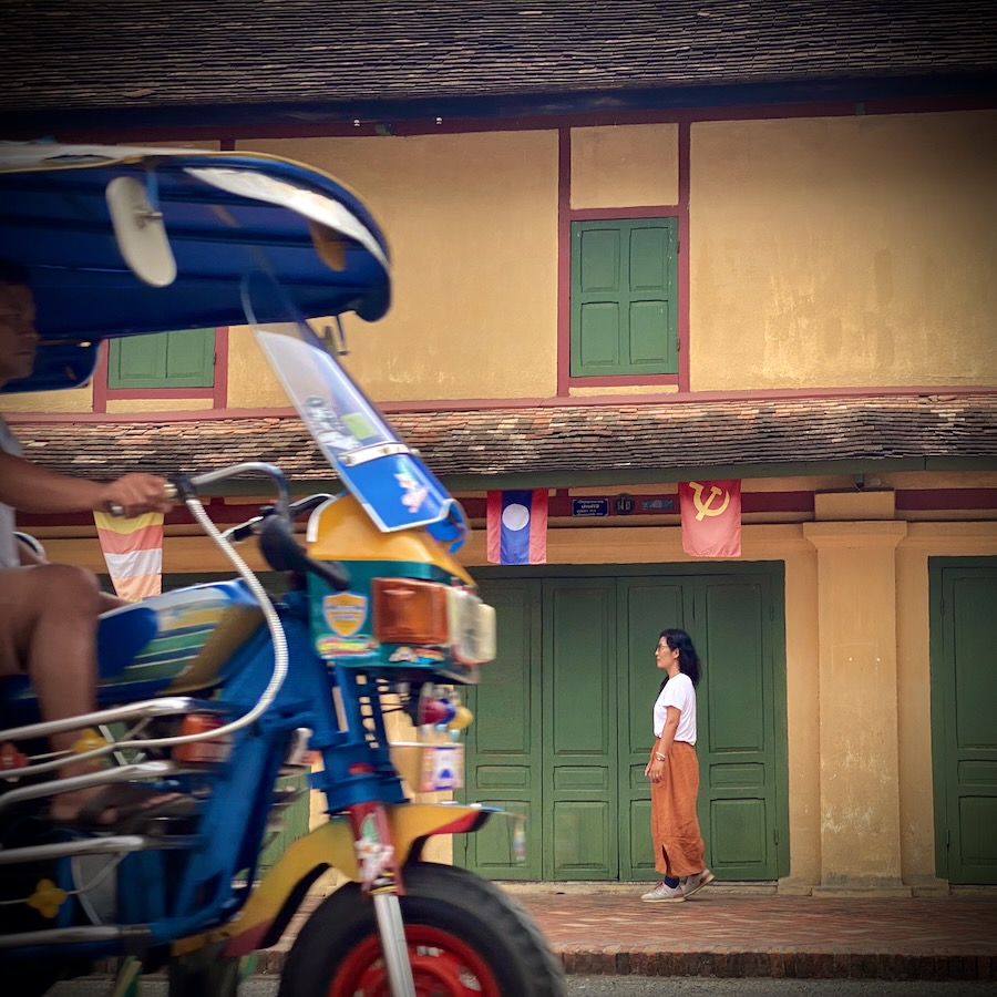 A tuk tuk passes a lady walking past colonial buildings in Luang Prabang in Laos