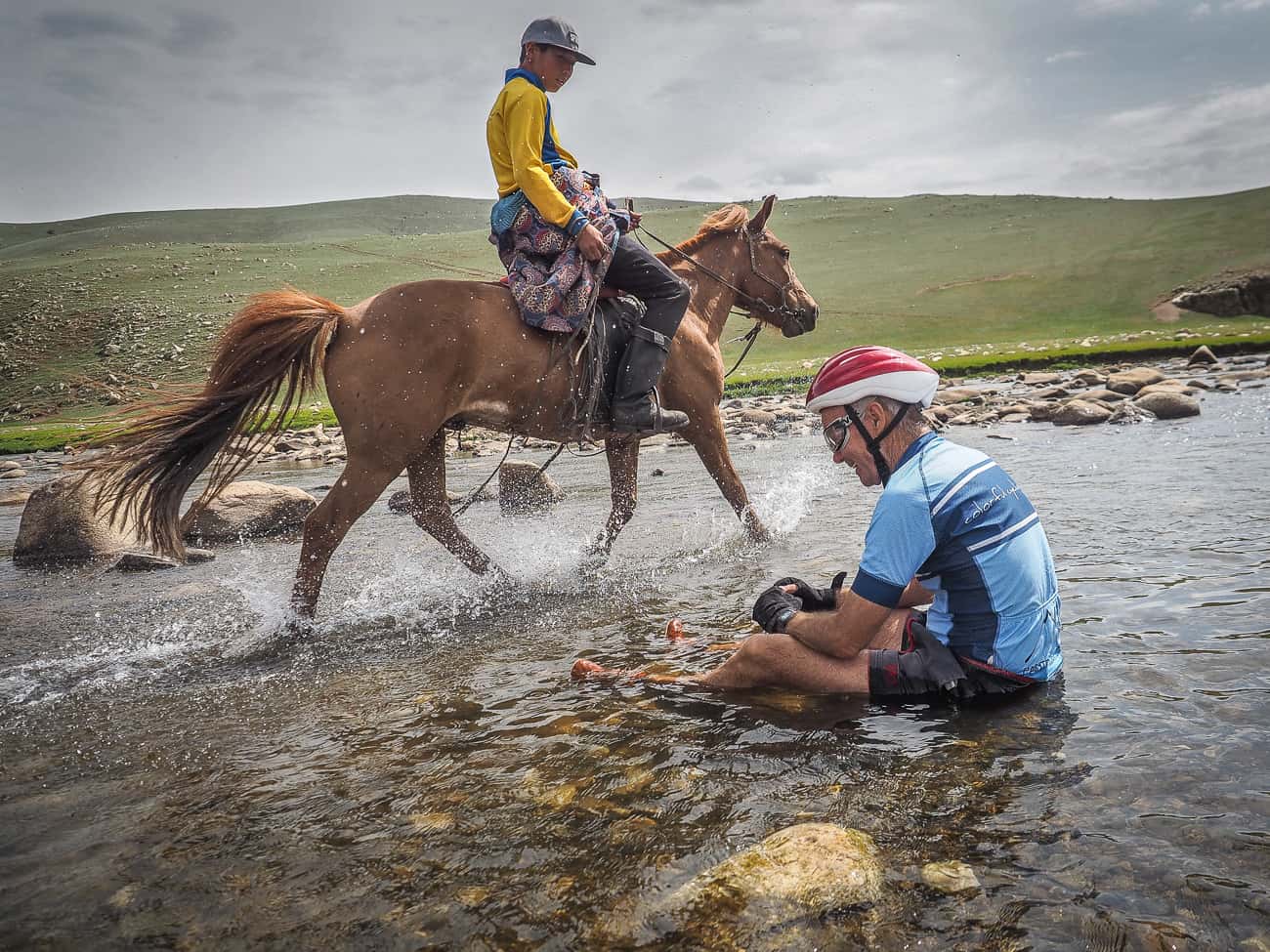 a cyclist in Mongolia sitting in a river as a horse rider passes by